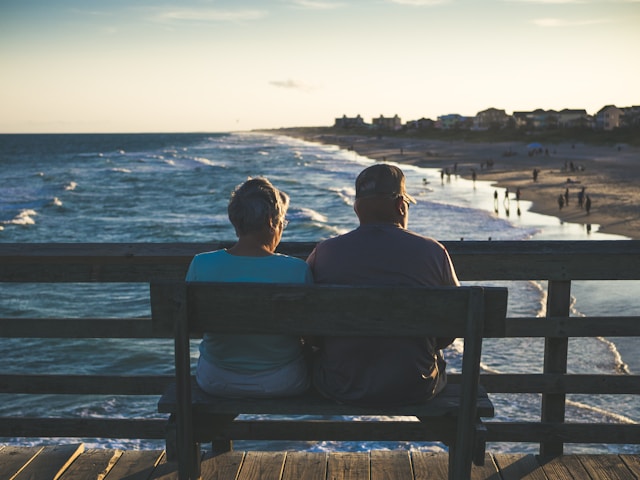 Afbeelding van 2 personen die op een bankje zitten en uitkijken over het strand en de zee.